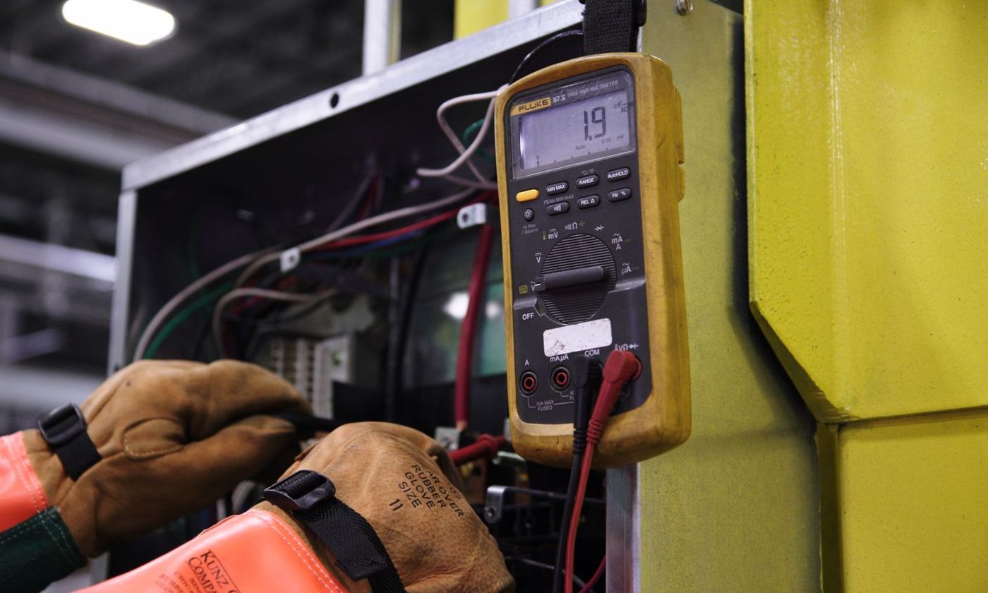 Technician in work gloves reading a digital multimeter at an open electrical panel